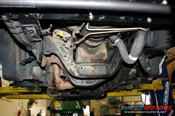Undercarriage view of vehicle suspension components and engine parts. Another vehicle is visible in the background