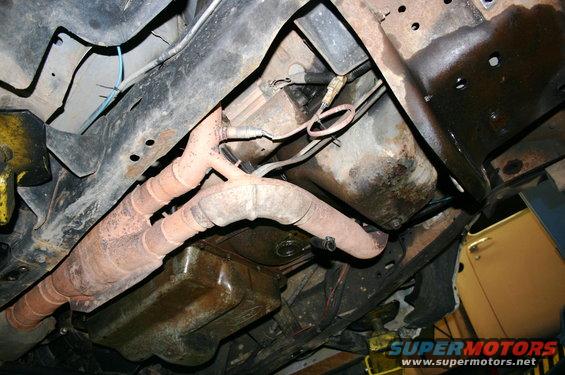 Underside view of a vehicle's exhaust system and frame. A yellow car is partially visible on the right
