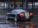 1965 Ford Mustang - A black Ford Mustang with gold racing stripes sits in a wet parking lot.