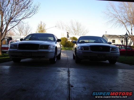 Two Ford Crown Victorias parked in a driveway, one white and one gray. The white one is on the left, gray on the right