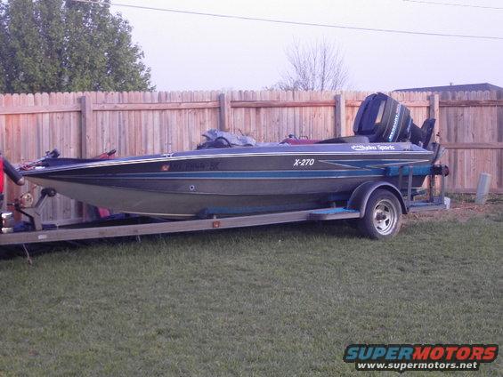 Boat with 'X-270' and 'Hydro Sports' decals on a trailer, parked on grass in front of a wooden fence
