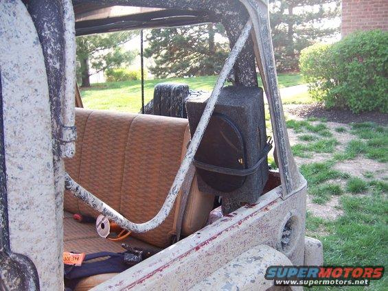 Jeep interior showing tan seats, roll bar, and a large speaker box mounted behind the passenger seat. Exterior is covered in white mud