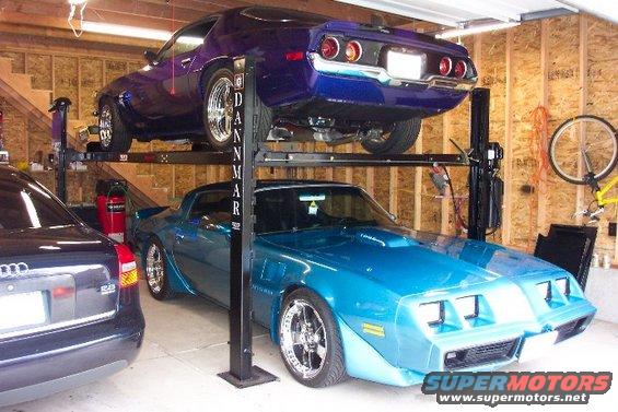 Blue 1979 Pontiac Trans Am on car lift, front 3/4 view. Purple Camaro above