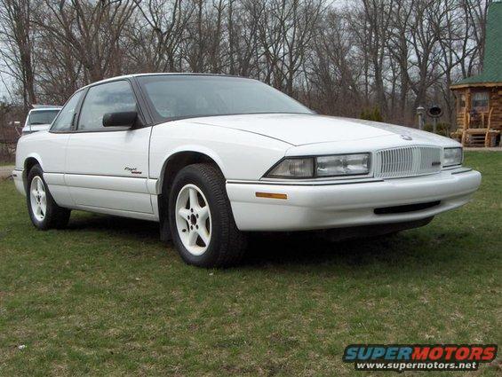 White coupe parked on grass with a log cabin in the background
