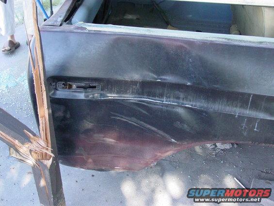 Damaged rear quarter panel of a dark vehicle, showing denting and a broken wooden plank