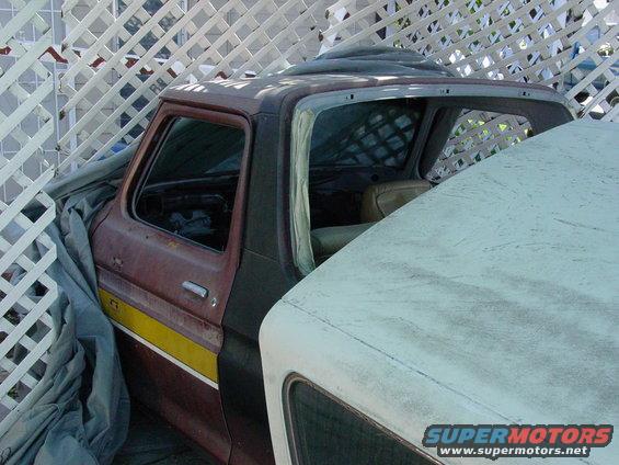 1979 Ford Bronco cab with yellow stripe, partially covered by tarp, next to another vehicle