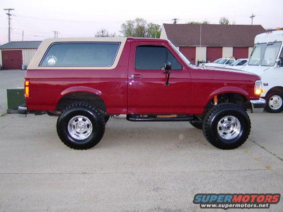 Red Ford Bronco with tan top, lifted suspension, chrome wheels, side view. Watermark 'SUPERMOTORS.NET' visible
