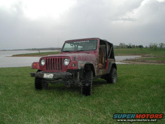 Red Jeep Wrangler parked on grass near a body of water. It has a soft top and the doors are off