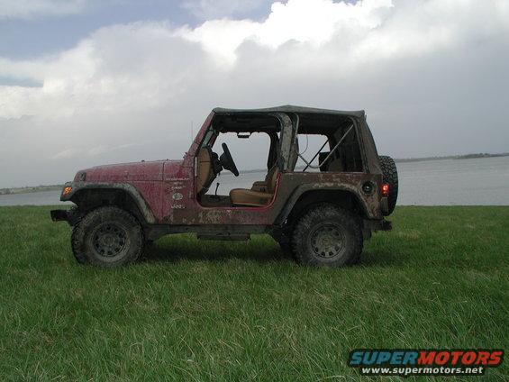 Red Jeep Wrangler with soft top removed, parked on a grassy field near a body of water