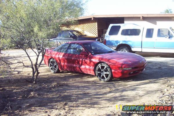 Red coupe with aftermarket wheels parked on dirt, a blue and white pickup truck behind it