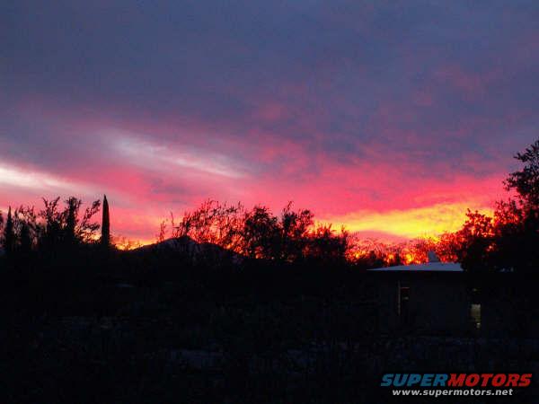 Desert sunset with colorful clouds and silhouetted trees and a building. Supermotors.net logo visible