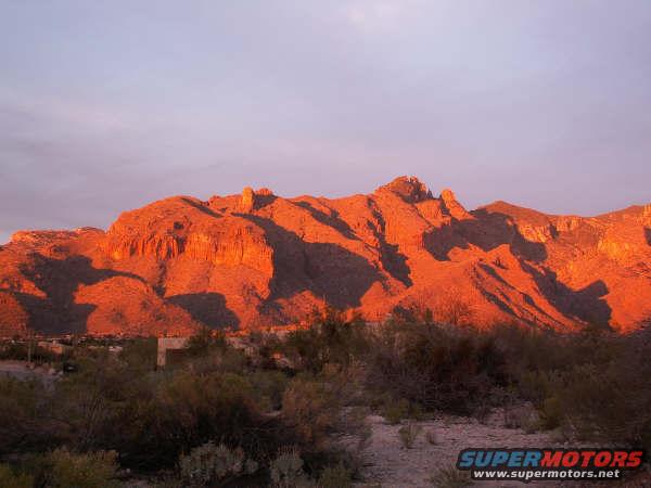 Mountains illuminated by sunset, with desert scrub in the foreground. A website URL is visible