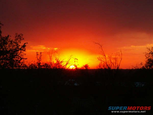 Sunset with vibrant orange and red sky over silhouetted trees and field. SUPERMOTORS.NET logo in lower right