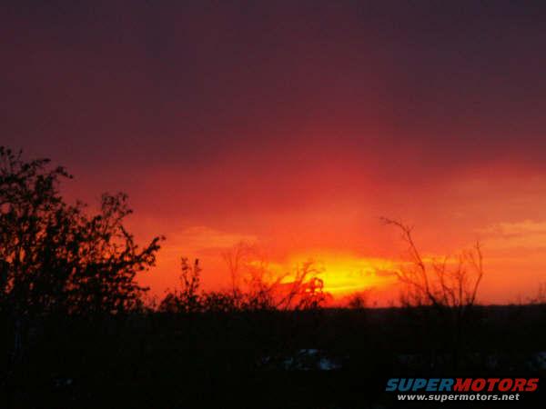Sunset sky with vibrant orange and red hues over a silhouetted field and trees. SUPERMOTORS.NET logo visible