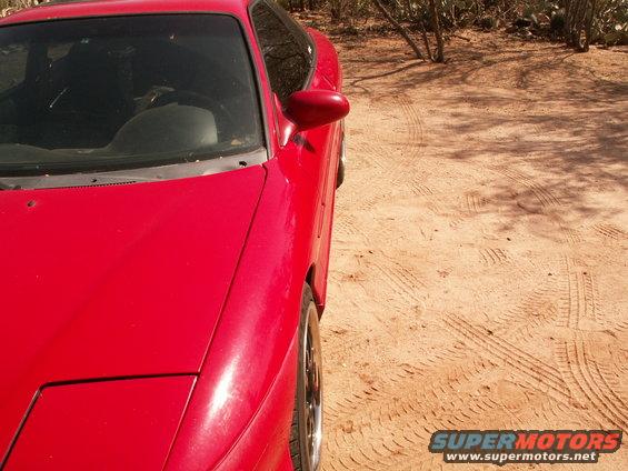 Red sports coupe parked on dirt with tire tracks. Side mirror and part of the hood are visible