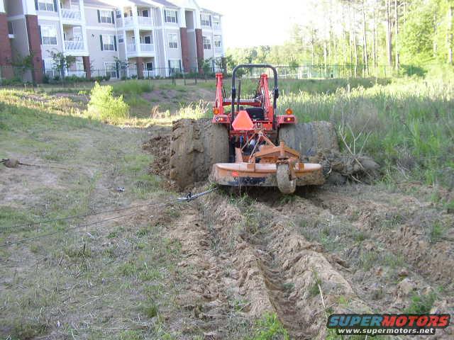 Orange tractor with mower attachment stuck in mud, pulling a cable