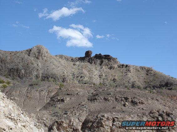 Rocky desert mountains under a blue sky with scattered clouds. Watermark reads SUPERMOTORS