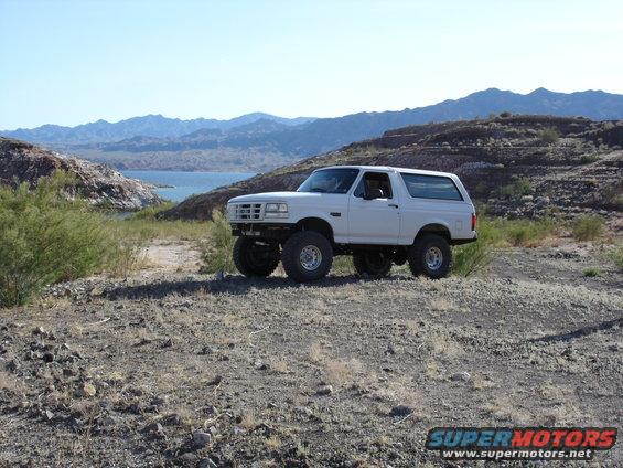 White 1996 Ford Bronco with large tires parked on a rocky hill overlooking a lake