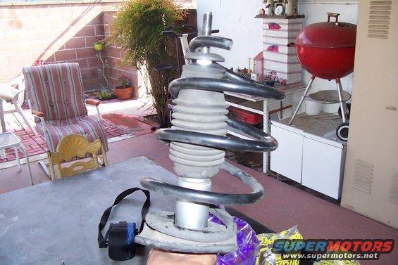 Car suspension strut assembly with a coil spring, sitting on a table outdoors. A red grill and chairs are in the background