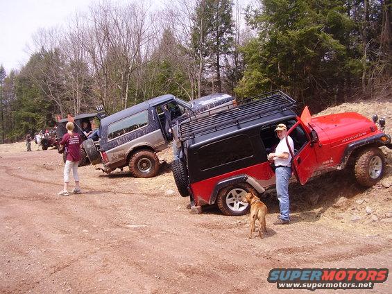 Red 2003 Jeep Wrangler Rubicon and a blue SUV on a dirt trail with trees in the background