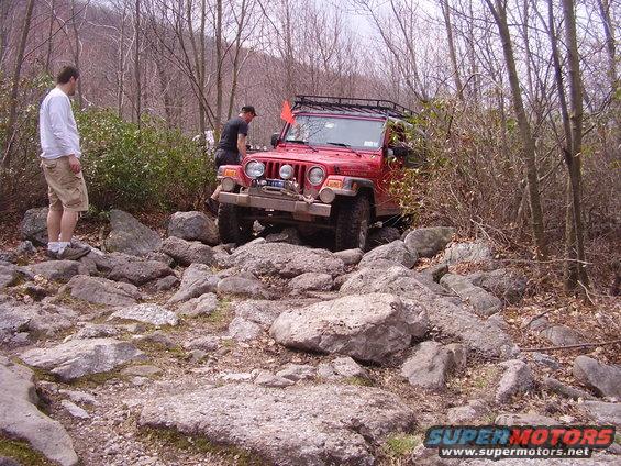 Red Jeep Wrangler navigating a rocky trail with two people nearby