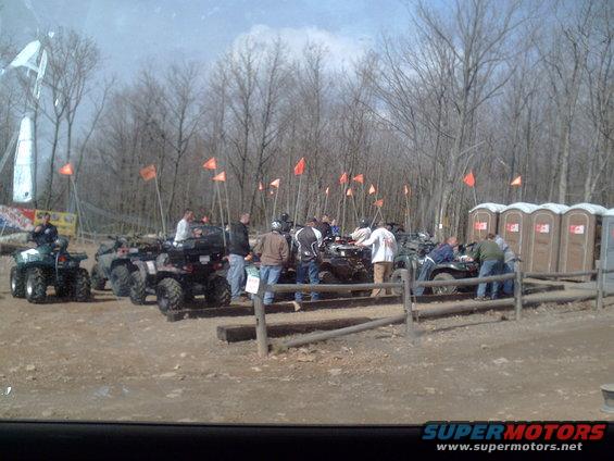 Group of people gathered around ATVs in a wooded area with portable toilets in the background