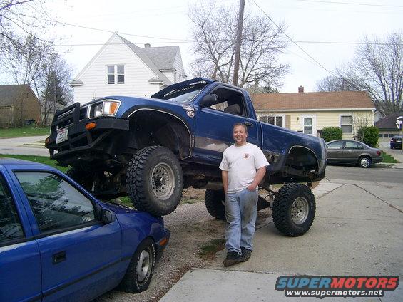 Lifted blue pickup truck with large tires, one front tire on top of a blue sedan. Man stands beside the truck