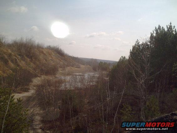 overcast sky with bright sun, dirt hills, trees, and a lake