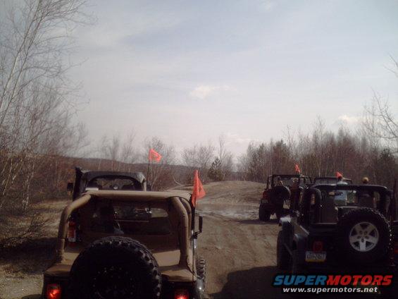 Several off-road vehicles, some with orange flags, drive on a dirt path through a wooded area