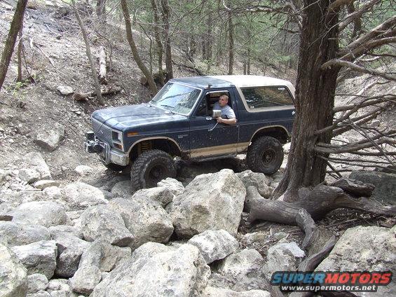 Dark blue Ford Bronco with a white top navigating a rocky trail, driver visible