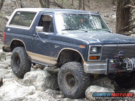 Dark blue Ford Bronco with a white top, lifted suspension, and large off-road tires, navigating rocky terrain