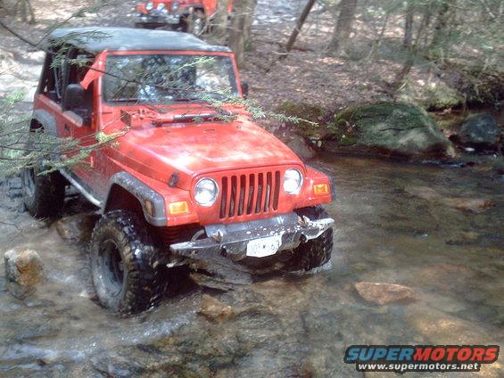 Red Jeep Wrangler driving through a rocky stream, another Jeep visible behind it