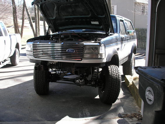 Ford Bronco, hood up, showing engine bay and front grille. Lifted suspension visible. Another white truck is parked to the left