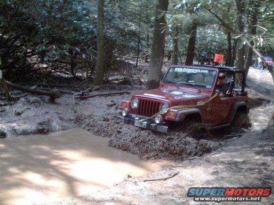 Red Jeep Wrangler stuck in a mud pit on a wooded trail. Another Jeep is visible in the background