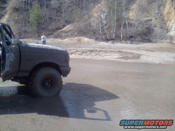 Muddy truck with door open, stuck in water. Off-road scene with hills in background