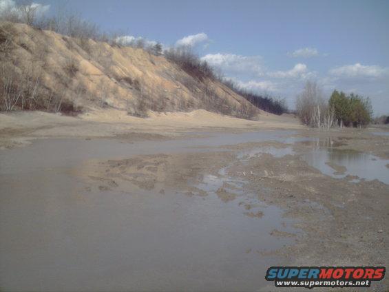 Muddy landscape with sand dune and trees under a blue sky with clouds