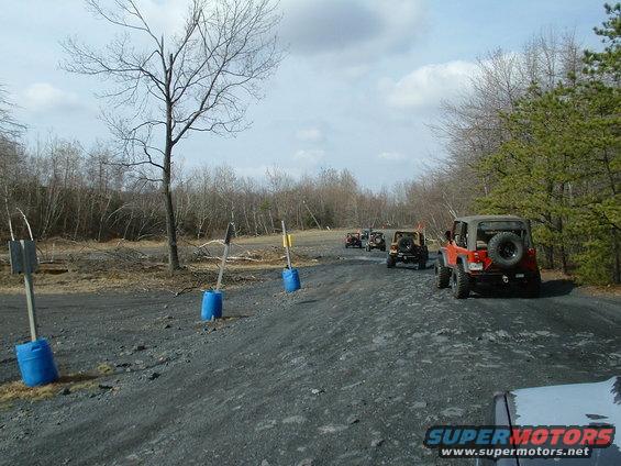 Several Jeeps driving in a line on a gravel road with trees and bare branches in the background
