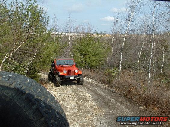 Orange Jeep Wrangler on a dirt road, with another vehicle's tire in the foreground