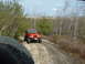 1991 Ford Bronco - An orange Jeep Wrangler drives on a rocky trail surrounded by trees and brush.