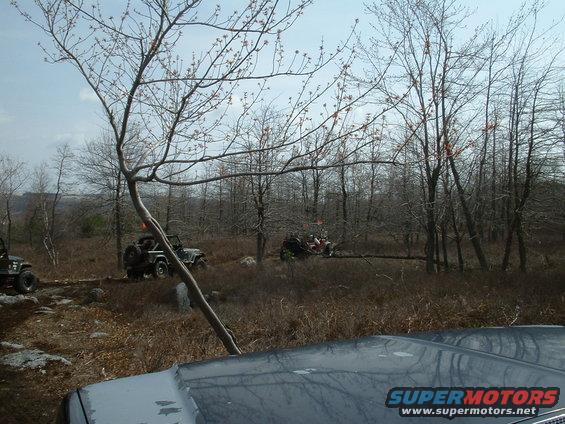 Several off-road vehicles navigate a rocky, wooded terrain under a cloudy sky