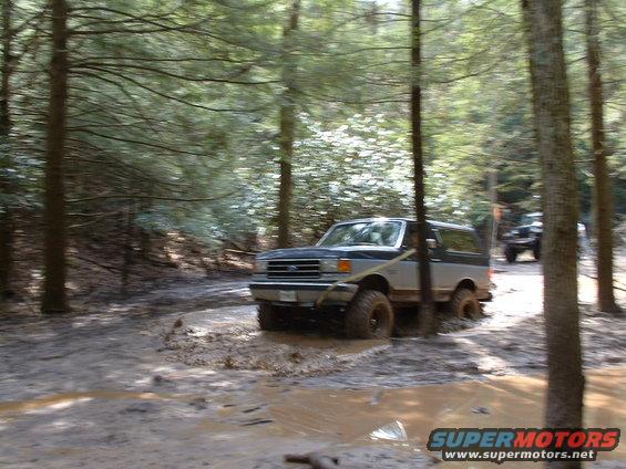 1991 Ford Bronco driving through a muddy trail in a forest, another vehicle visible in the background