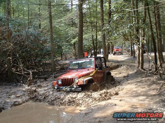 Red Jeep Wrangler stuck in mud on wooded trail; "SUPERMOTORS.NET" logo visible