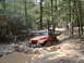 1991 Ford Bronco - A red Jeep Wrangler struggles through a muddy off-road trail in a forest, with other vehicles visible in the background.