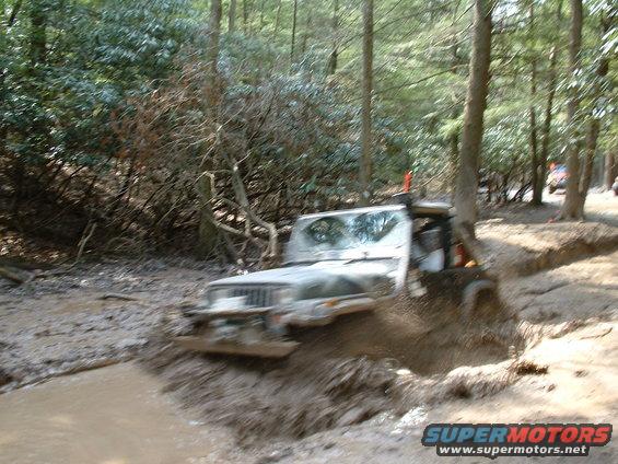 Jeep Wrangler going through a muddy trail in the woods. Another vehicle is visible in the background