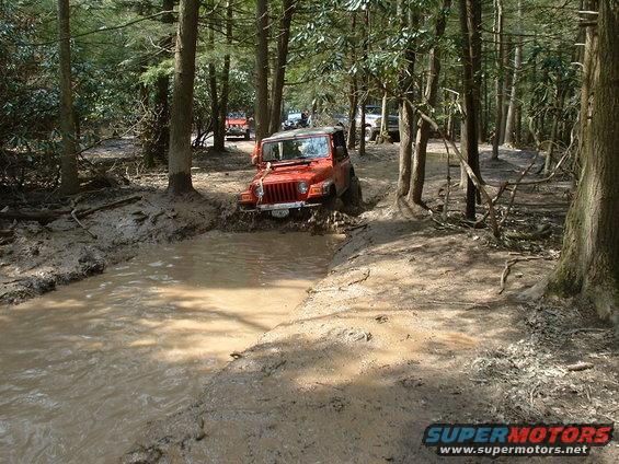 Orange Jeep Wrangler navigating a muddy trail with other vehicles behind it