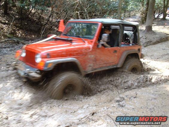 Orange Jeep Wrangler driving through mud, front-left wheel deep in muck