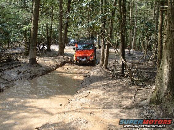 Orange Jeep Wrangler driving through a muddy trail in a forest; another vehicle is visible behind it