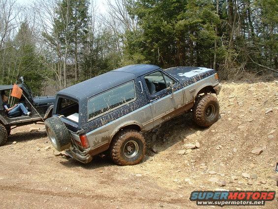 Mud-splattered blue and silver Ford Bronco on a dirt hill with a spare tire on the back