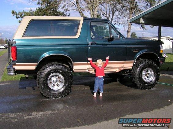 Green 1995 Ford Bronco with tan trim, lifted suspension, and large tires, a child stands in front of it