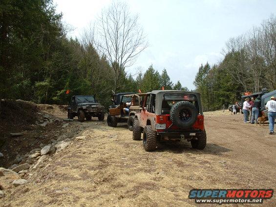 Several Jeeps are parked and driving on a dirt trail in a wooded area. One red Jeep is in the foreground